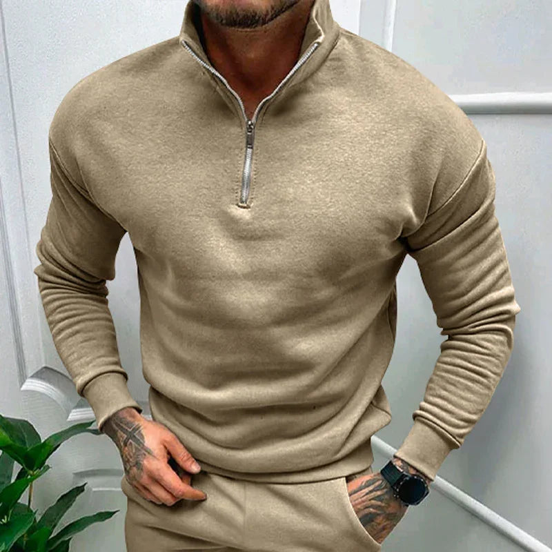 Man wearing beige quarter-zip sweatshirt and matching pants with tattooed hands and black watch