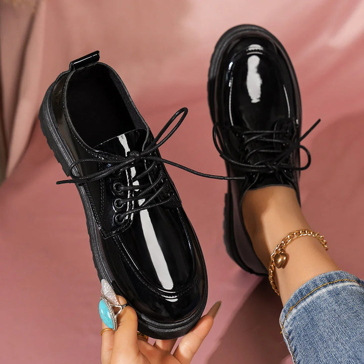 Glossy black patent leather lace-up shoes worn with blue jeans and gold anklet on pink background