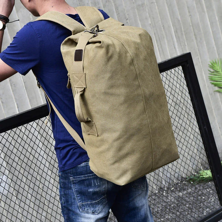 Man wearing a large khaki canvas backpack with adjustable straps, standing by a metal fence