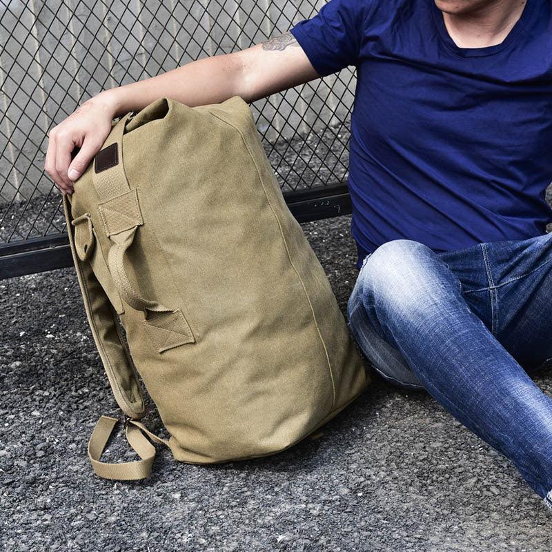 Person in blue shirt and jeans sitting on pavement holding a large khaki canvas backpack beside a chain-link fence