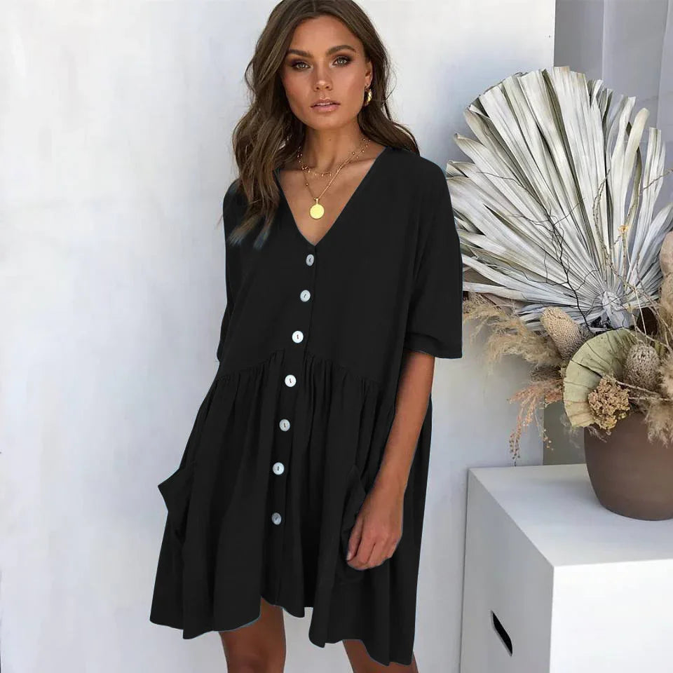 Woman wearing black button-down loose dress with pockets, standing next to modern vase with dried palm leaves