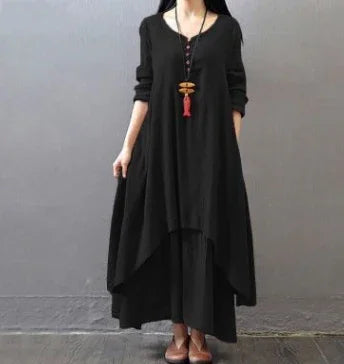 Woman wearing a loose black asymmetrical dress with long sleeves and a long pendant necklace against a gray background