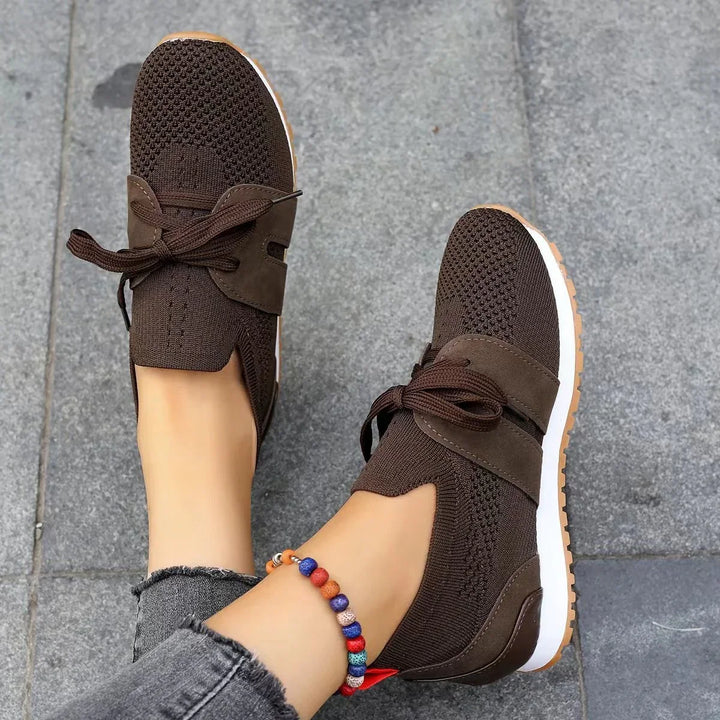 Brown knit fabric sneakers with laces and white soles worn with gray frayed jeans on stone pavement