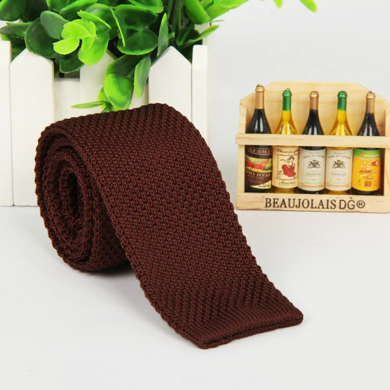 Rolled dark brown knitted tie displayed on white surface with green plant and wine bottle decor