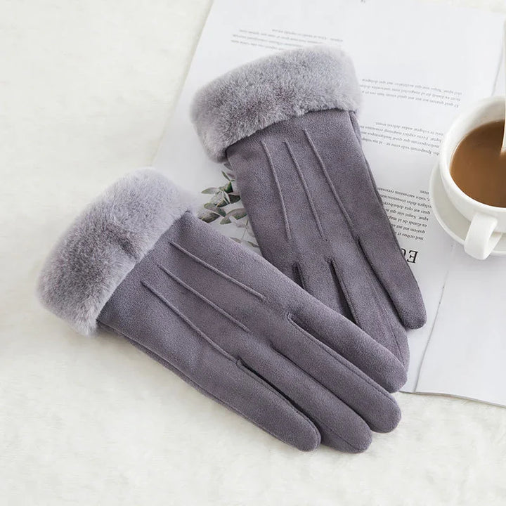 Gray winter gloves with soft fur cuffs on a white surface next to a cup of coffee and an open book
