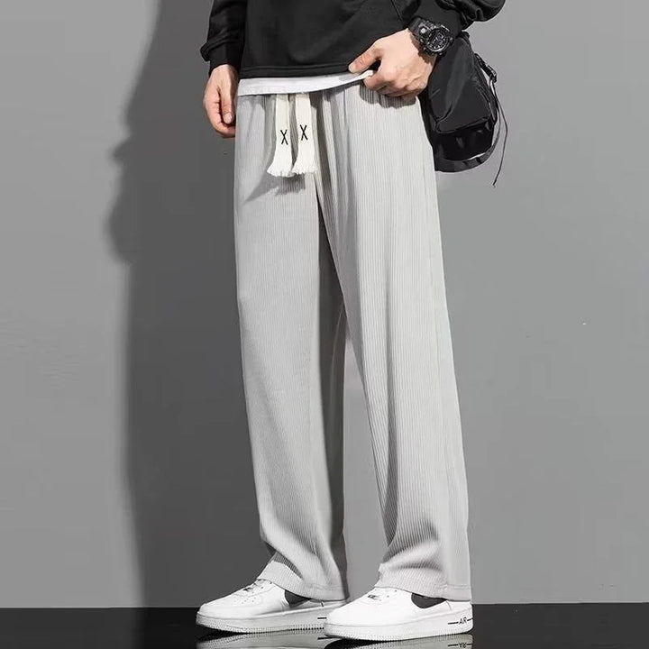 Man wearing light gray ribbed wide-leg pants with white drawstrings and white sneakers against gray background