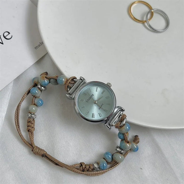 Silver quartz wristwatch with braided cord strap and blue beads, placed on white surface near gold and silver rings