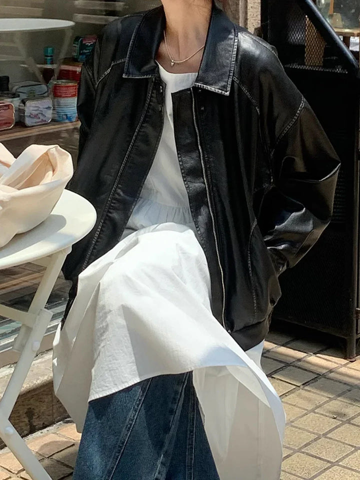 Woman sitting outdoors wearing a black leather jacket, white dress, and blue jeans next to a white table
