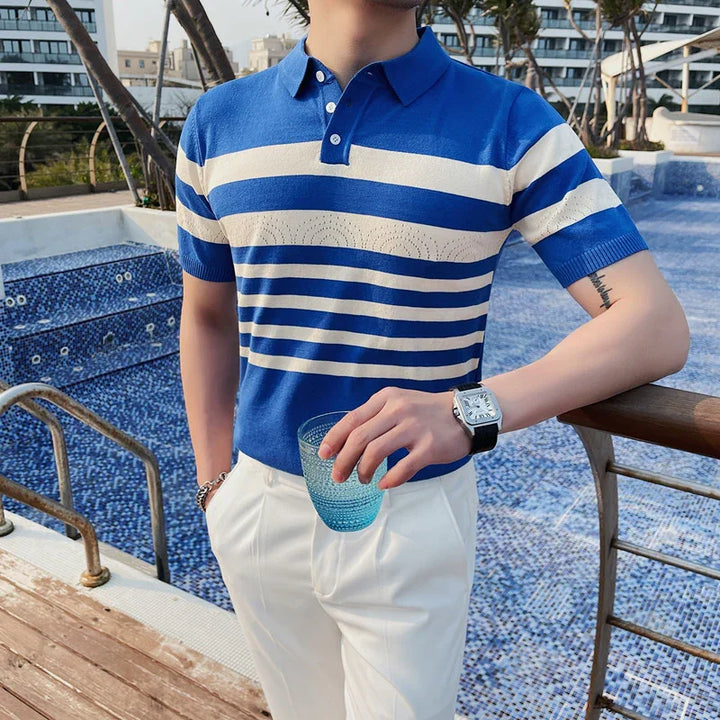 Man wearing blue and white striped polo shirt and white pants holding blue glass by poolside
