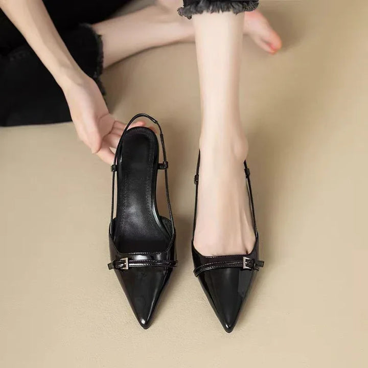 Close-up of black patent leather pointed-toe slingback heels worn by model sitting on beige floor
