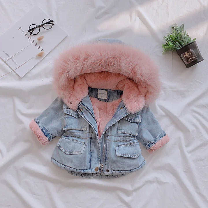 Baby denim jacket with pink faux fur lining and hood on white fabric with glasses and plant