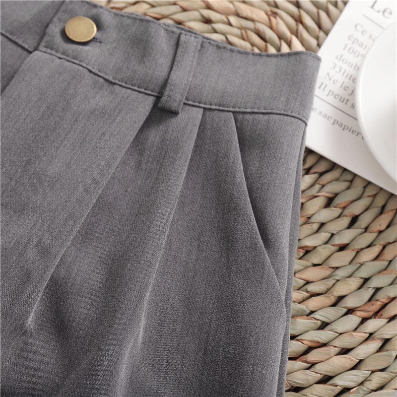 Close-up of gray high-waisted pants with pleats and brass button on woven mat background
