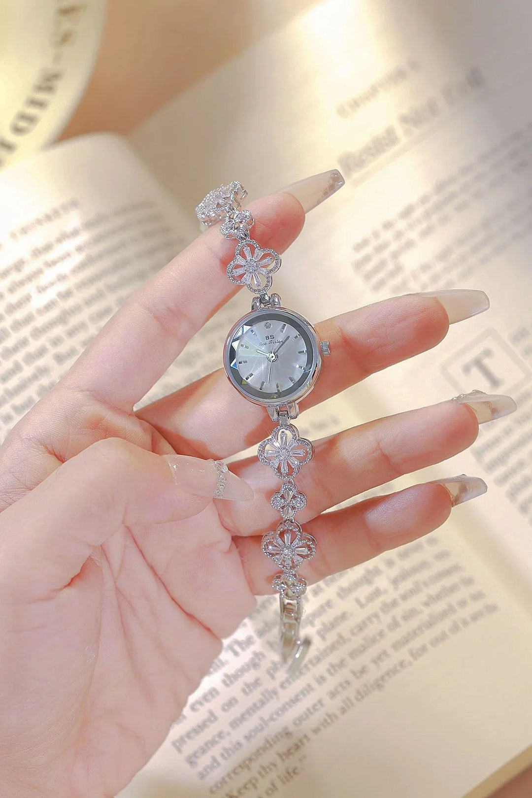 Hand holding elegant silver floral bracelet watch with clear long nails over open book