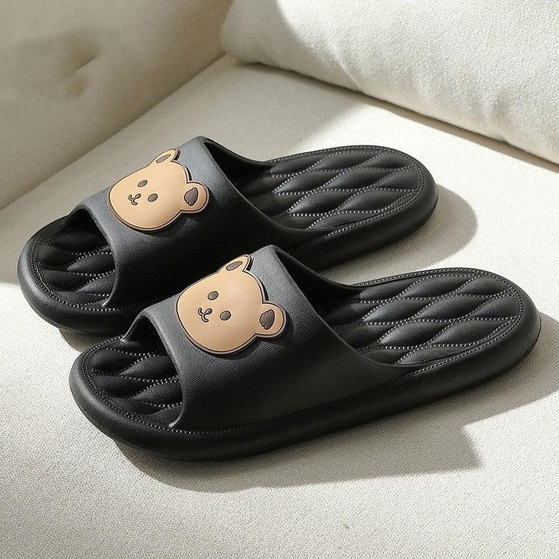 Black quilted slide sandals with cute tan bear face decoration on top, placed on white fabric surface