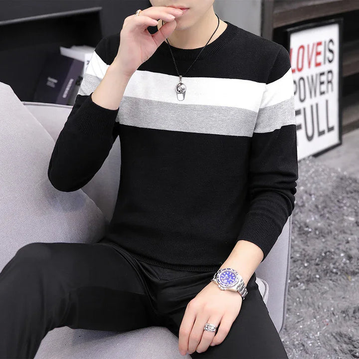Man wearing black sweater with white and gray stripes, black pants, silver watch, and necklace sitting on gray sofa