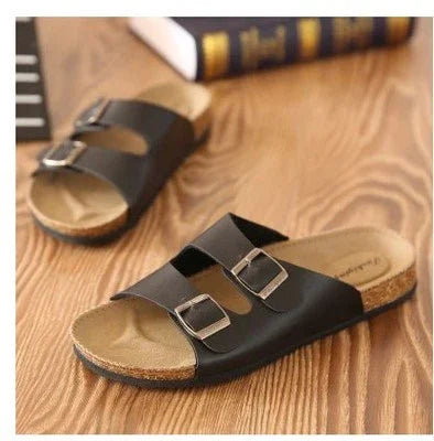 Black double buckle slide sandals with cork footbed on wooden floor near a book