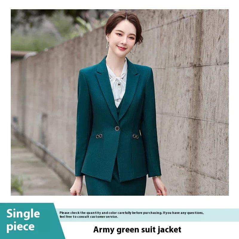 woman wearing army green suit jacket with silver eyelet details, white blouse, standing by concrete wall