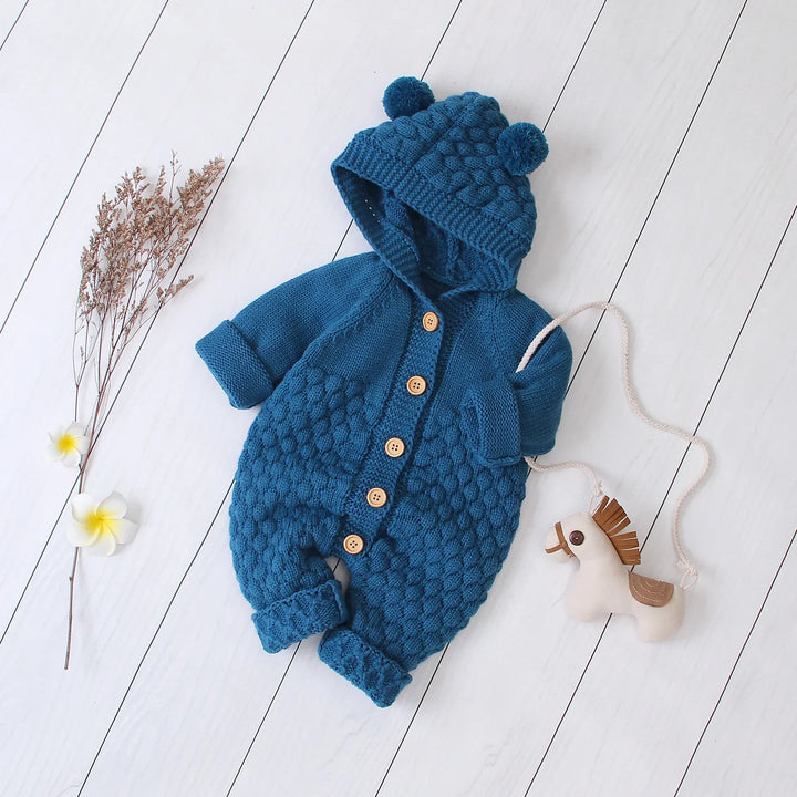 Blue knitted baby romper with hood and pom-poms, wooden buttons, on white wooden floor with dried flowers and toy horse