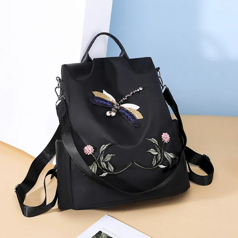 Black backpack with jeweled dragonfly and floral embroidery, adjustable straps, on beige and blue background