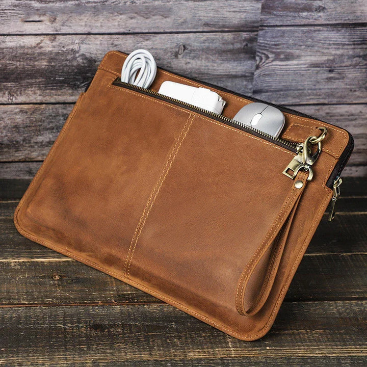 brown leather tech organizer pouch with wrist strap, containing white charger and silver mouse, on wooden surface
