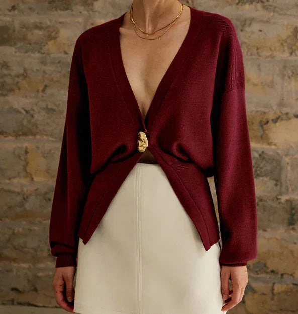 Woman wearing deep burgundy V-neck cardigan with gold clasp and white skirt against stone wall