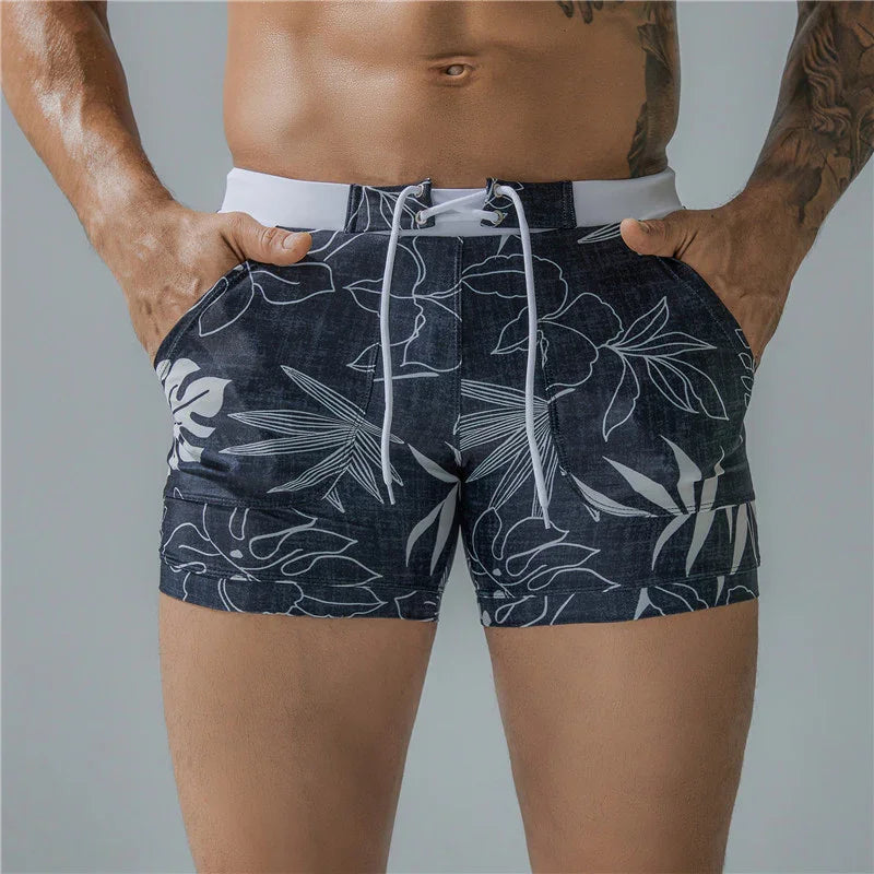 Close-up of man's torso wearing dark floral print swim trunks with white drawstrings, hands in pockets