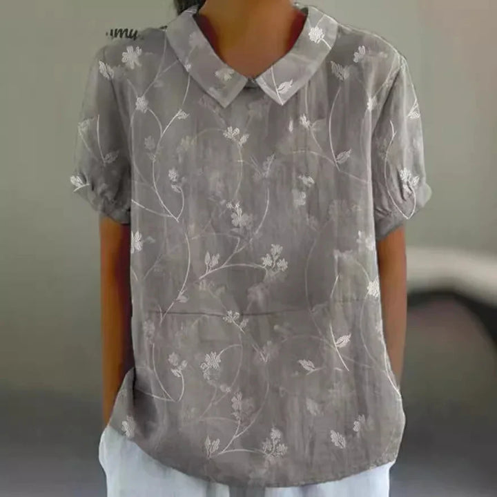 Gray short-sleeve blouse with white floral embroidery and a pointed collar worn by a person
