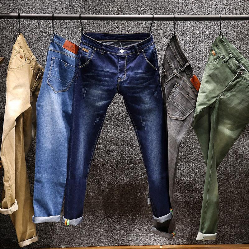 Five pairs of men's casual pants in beige, blue, dark blue, gray, and green hanging on a clothing rack against a textured gray wall