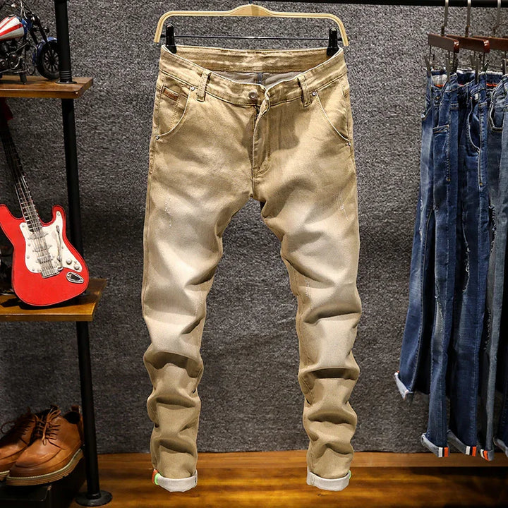 Beige slim-fit men's jeans hanging on wooden hanger in retail store with blue jeans and accessories