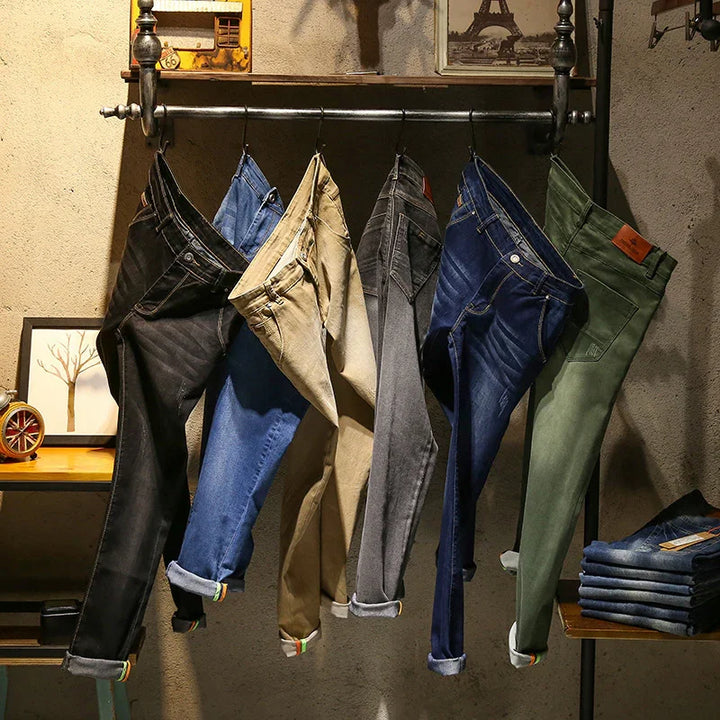 Six pairs of jeans in black, blue, beige, grey, dark blue, and green hanging on a metal rack in a rustic room