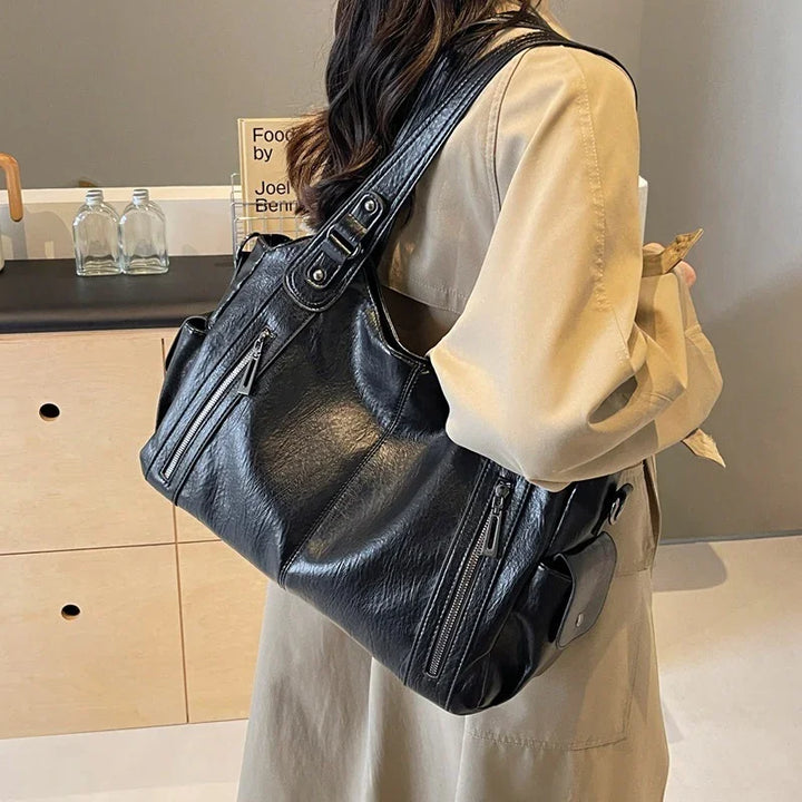 Woman wearing beige coat carrying stylish black leather shoulder bag with zipper pockets indoors
