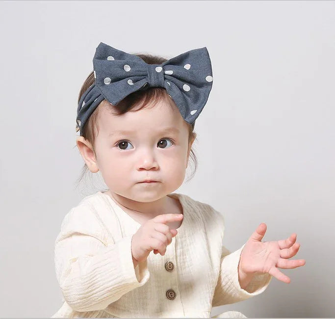Toddler wearing cream buttoned outfit and blue polka dot bow headband on gray background