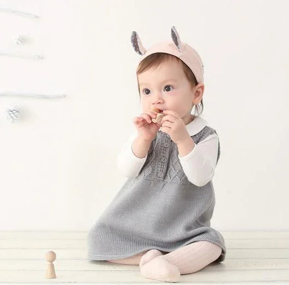 Baby wearing gray knit dress and cute animal ear headband sitting on wooden floor with wooden toy
