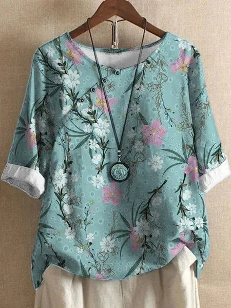 Light blue floral blouse with pink and white flowers, rolled sleeves, paired with beige pants and round pendant necklace