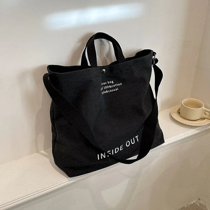 Black canvas tote bag with white text "INSIDE OUT" on a white shelf next to a beige cup and saucer
