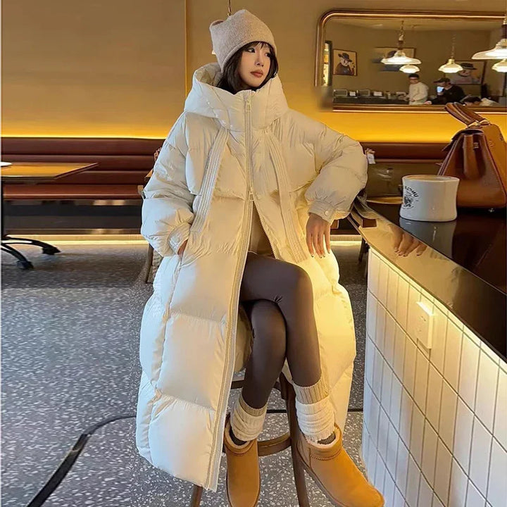 Woman in beige long puffer coat, beanie, and tan boots sitting on stool in cozy café