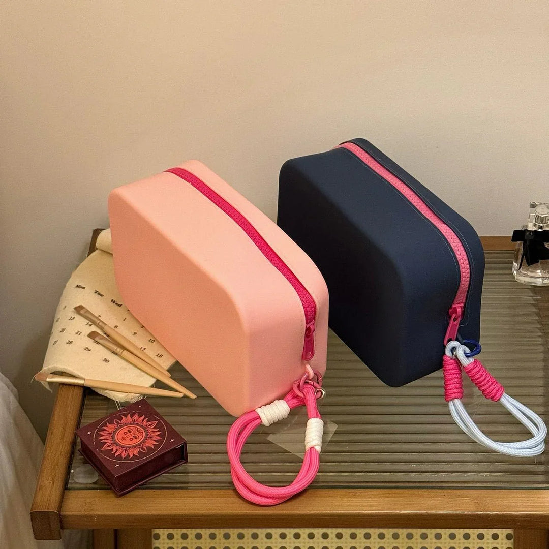 Two rectangular silicone makeup bags in pink and navy with contrasting pink zippers and wrist straps on a glass wooden table