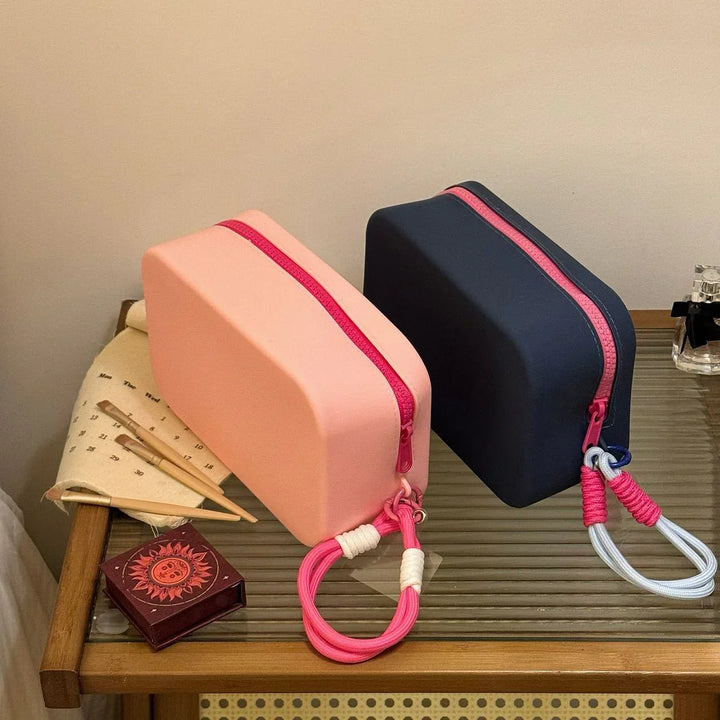Two rectangular silicone makeup bags in pink and navy with contrasting pink zippers and wrist straps on a glass wooden table