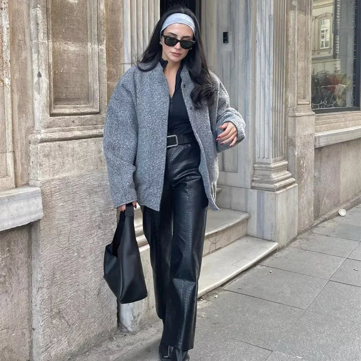 Woman in gray oversized jacket and black leather pants walking on city sidewalk carrying black handbag