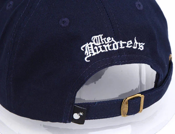Navy blue adjustable cap with embroidered The Hundreds logo on back and brass buckle strap