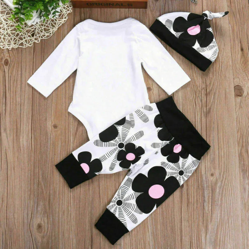 White baby bodysuit with black and pink floral pants and matching hat on wooden background