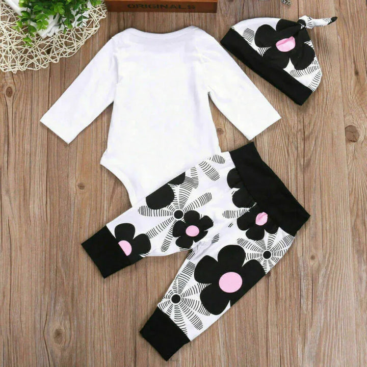 White baby bodysuit with black and pink floral pants and matching hat on wooden background