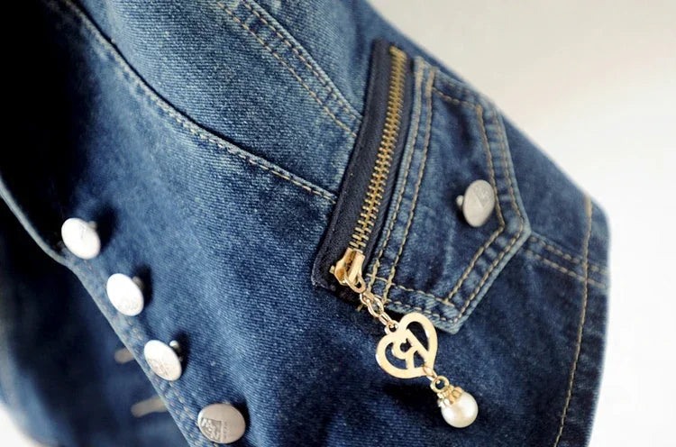 Close-up of blue denim jacket sleeve with silver buttons and gold zipper pull featuring heart and pearl charm