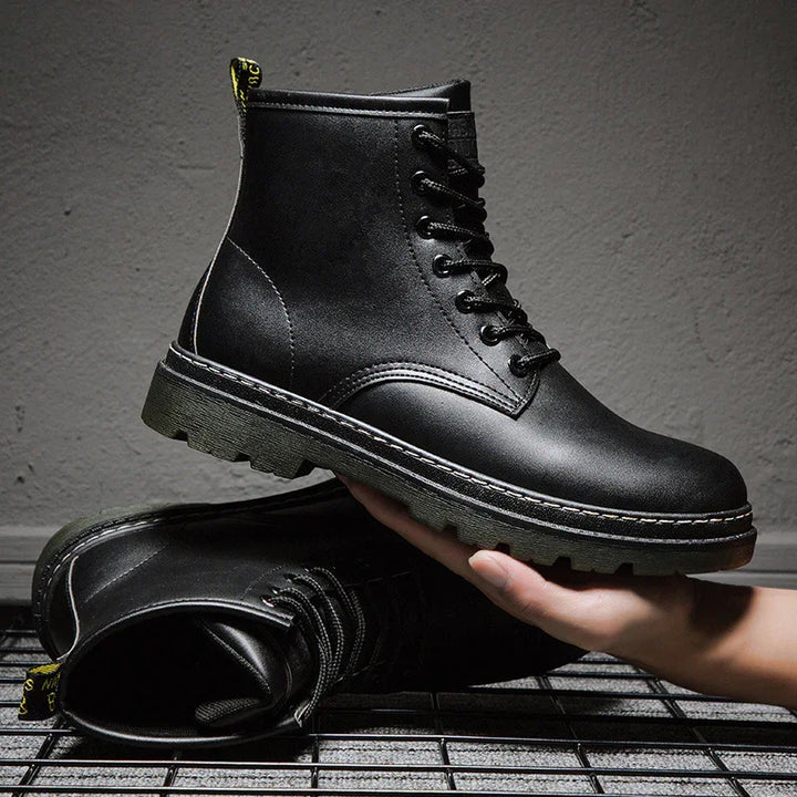 Black leather combat boots with thick sole, hand holding one boot, industrial background