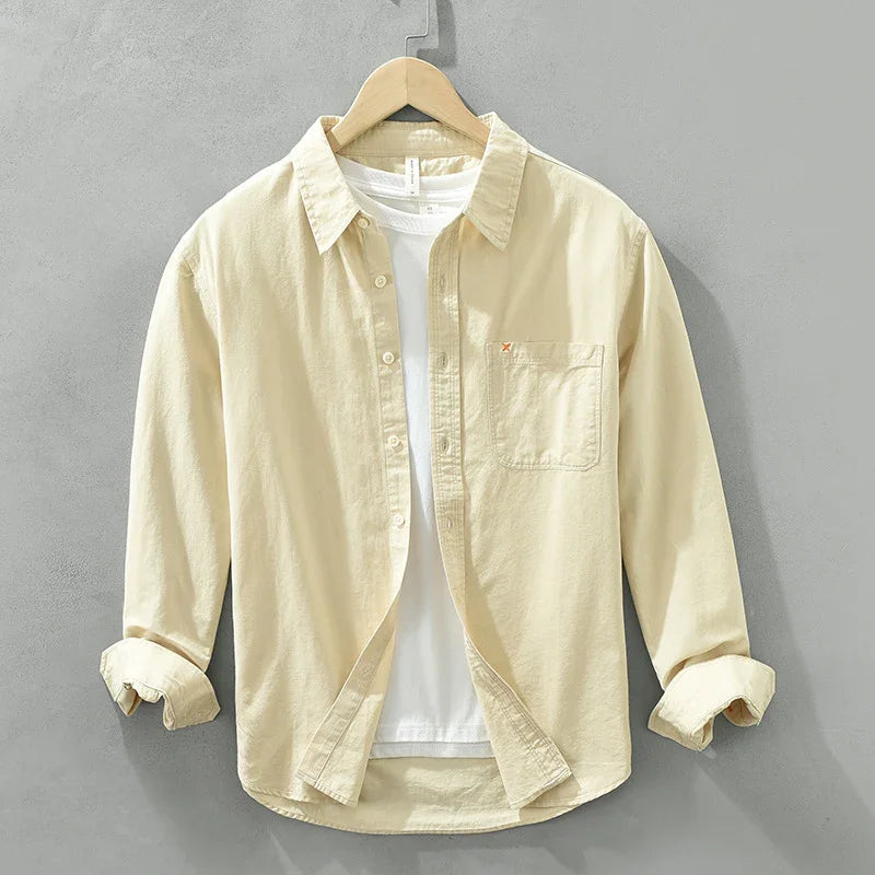 Beige button-up shirt with rolled sleeves over white t-shirt on wooden hanger against gray wall