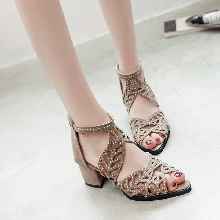 Woman wearing beige studded block heel sandals with intricate cut-out design on gray floor