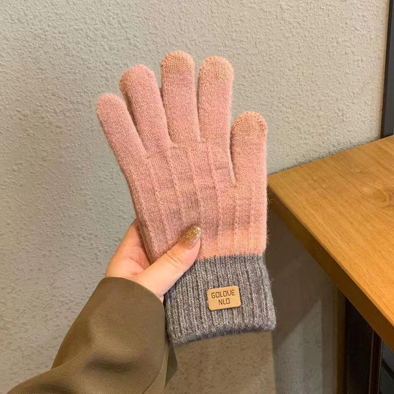 Hand holding pink and gray knitted gloves with a small beige label against a textured wall and wooden surface