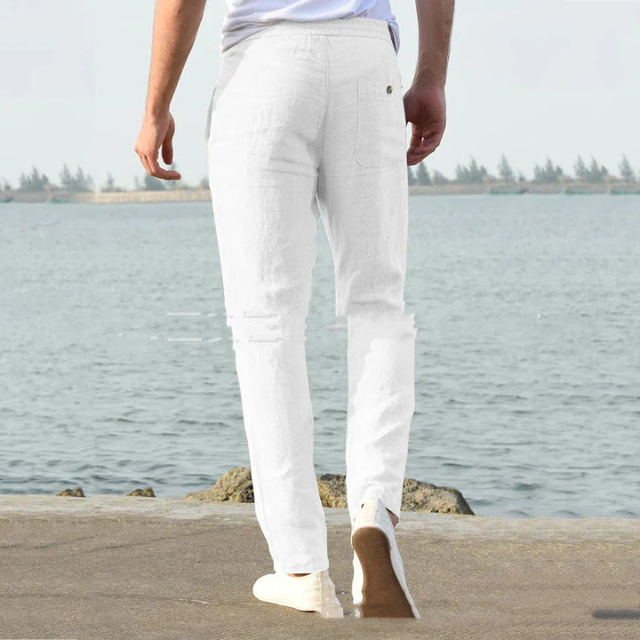 Man wearing white linen pants and casual shoes walking near a waterfront with calm water and distant trees