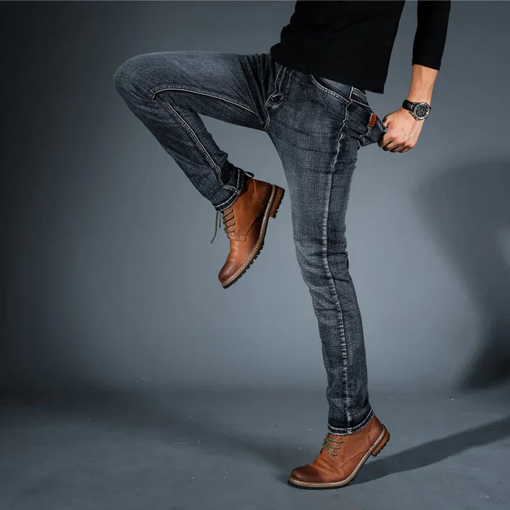 Man wearing slim fit dark blue jeans and brown leather boots against gray background
