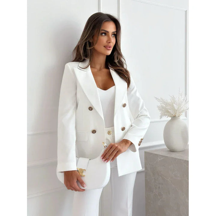 Woman in white tailored blazer and pants holding a white handbag in a minimalist room